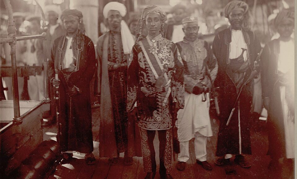 Au centre de l’image, le jeune prince Salim bin Abdallah de Ndzuani apparaît richement décoré. Il arbore quatre croix de l’Étoile de Ndzuani sur le côté gauche de sa veste ainsi qu’une grand-croix portée au cou. Il pose, en 1883, sur le pont d’un navire aux côtés d’une délégation de cinq dignitaires. © Smithsonian Institution