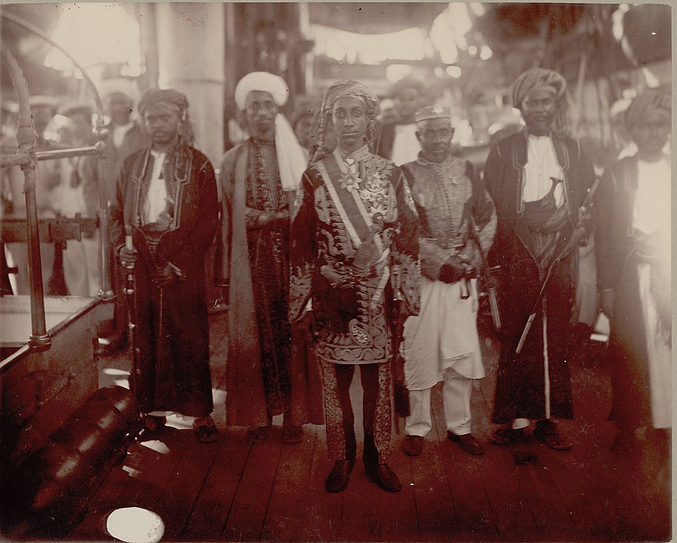 Au centre de l’image, le jeune prince Salim bin Abdallah de Ndzuani apparaît richement décoré. Il arbore quatre croix de l’Étoile de Ndzuani sur le côté gauche de sa veste ainsi qu’une grand-croix portée au cou. Il pose, en 1883, sur le pont d’un navire aux côtés d’une délégation de cinq dignitaires. © Smithsonian Institution